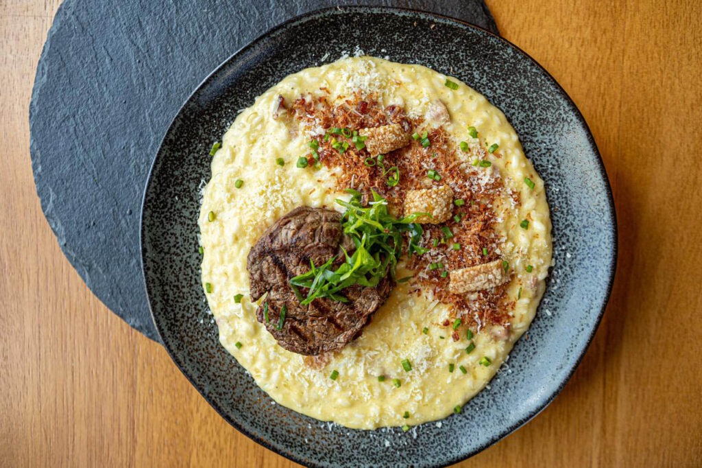 A imagem mostra um prato de comida gourmet, composto por um risoto cremoso coberto com pedaços crocantes de torresmo, cebolinha picada e queijo ralado. Ao centro, há um medalhão de carne decorado com folhas verdes. O prato está servido em um prato fundo de cor preta, sobre uma mesa de madeira clara.