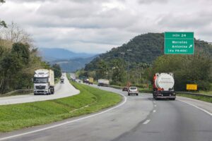 A imagem mostra uma estrada duplicada com tráfego moderado de caminhões e carros, cercada por vegetação densa e montanhas ao fundo sob um céu nublado. No lado direito da estrada, há uma placa verde indicando a "Saída 24" para Morretes e Antonina, via PR-408.