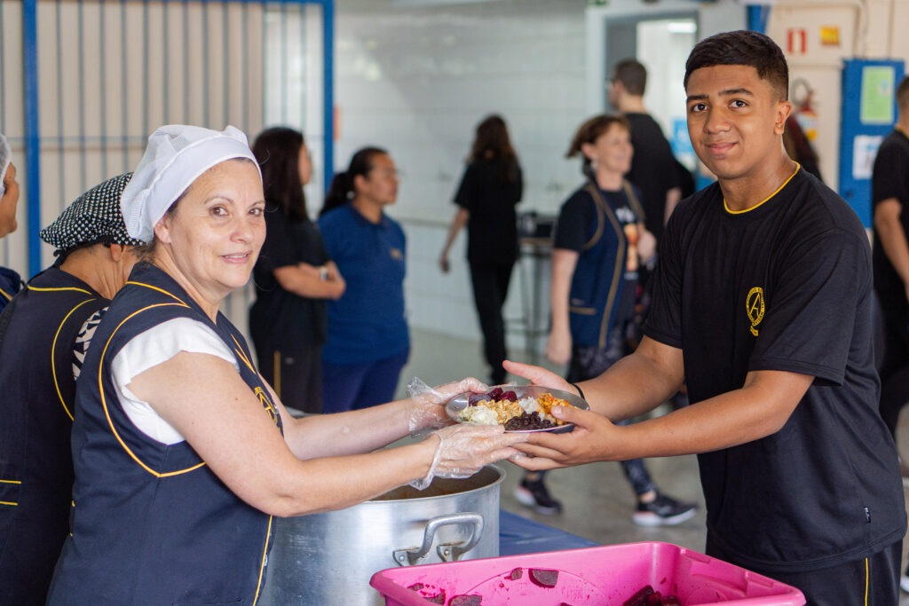 Á esquerda, uma pessoa vestindo um colete azul e toca de cabelo entrega um prato de comida para um jovem à direita que recebe com ambas as mãos.