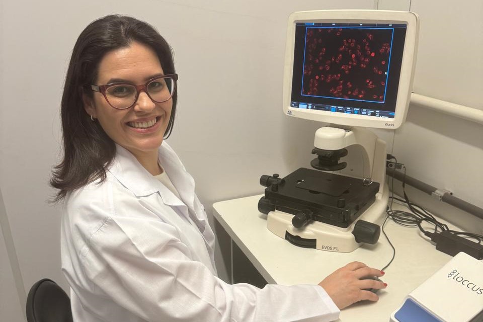 A imagem mostra uma pesquisadora em um laboratório. Ela está sorrindo para a câmera, vestindo um jaleco branco e óculos, e está sentada em frente a um microscópio digital acoplado a uma tela de computador. Na tela, é possível ver uma imagem ampliada de células vermelhas, que parecem estar sendo analisadas. A pesquisadora está manipulando o equipamento com uma das mãos. O ambiente parece limpo e bem iluminado, típico de um laboratório científico, com cabos e instrumentos ao redor da mesa. A pesquisa parece estar relacionada ao estudo de células, possivelmente no contexto de tratamentos para o câncer de mama.