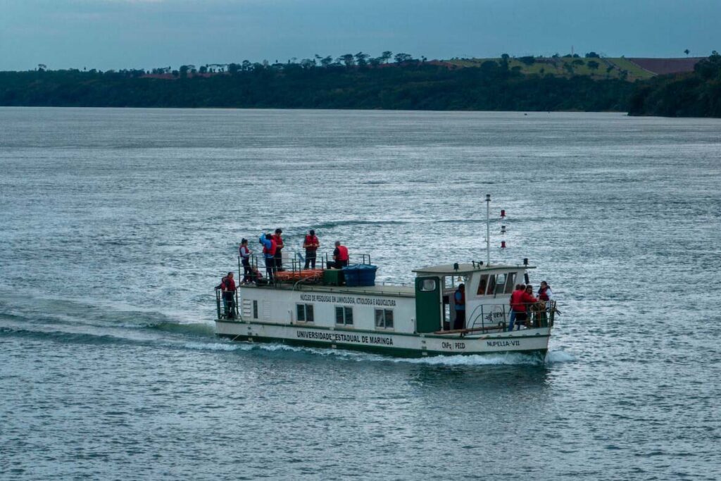 A imagem mostra um barco de pesquisa navegando em um rio largo, com algumas pessoas a bordo usando coletes salva-vidas vermelhos. O barco é da Universidade Estadual de Maringá (UEM) e está identificado com a inscrição "Núcleo de Pesquisas em Limnologia, Ictiologia e Aquicultura" em sua lateral. O nome do barco é "NUPELIA VII". Várias pessoas estão na parte superior e na frente do barco, provavelmente participando de atividades de pesquisa relacionadas à biodiversidade aquática. Ao fundo, vemos a margem do rio coberta por vegetação, com colinas verdes que se estendem ao longo da paisagem, e o céu parece nublado, sugerindo um dia parcialmente encoberto.