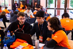 A imagem mostra três jovens universitários sentados em uma mesa, todos focados em uma conversa enquanto olham para o laptop aberto à sua frente. Eles estão participando do Hackathon Copel 2024, um evento de programação e inovação. O ambiente é movimentado, com outras pessoas ao fundo, também concentradas em suas tarefas. O jovem à esquerda, de barba e óculos, gesticula enquanto parece explicar algo aos outros dois, que estão atentos. Eles estão usando crachás e há mochilas e garrafas de água na mesa. Ao fundo, outras pessoas vestem camisetas laranjas com a inscrição "Hackathon Copel". O local tem grandes janelas, permitindo a entrada de luz natural, sugerindo um espaço bem iluminado e arejado.