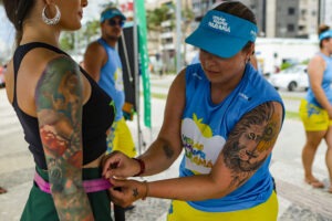 A imagem mostra uma pessoa medindo a cintura de outra pessoa com uma fita métrica. Ambas estão em um ambiente ao ar livre, possivelmente em um evento esportivo ou de saúde. A pessoa que está realizando a medição veste um uniforme azul com a logomarca do programa "Verão Maior Paraná" e usa um boné azul. Ela tem tatuagens visíveis nos braços, incluindo um leão com um girassol em um dos braços. A pessoa sendo medida também possui tatuagens grandes no braço, incluindo uma imagem de uma mulher estilizada. Ao fundo, outras pessoas participam do evento, e a cena parece se passar em uma área urbana à beira-mar, com algumas construções e árvores ao redor.