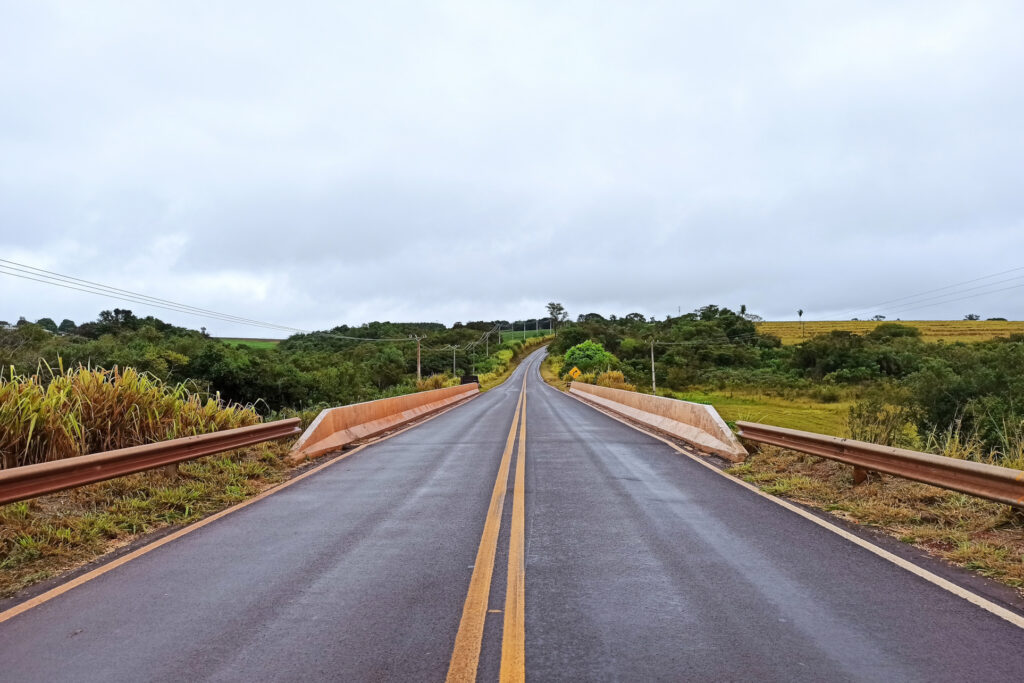 A imagem mostra uma estrada asfaltada, vista de frente, se estendendo em linha reta sobre uma ponte. A estrada possui uma faixa amarela dupla contínua no centro e guarda-corpos metálicos nas laterais, em um cenário rural com vegetação verde dos dois lados. Ao fundo, o caminho segue em direção a colinas levemente elevadas, com árvores e plantações ao redor. O céu está nublado, criando uma atmosfera cinzenta e úmida, indicando que pode ter chovido recentemente, já que o asfalto está levemente brilhante.