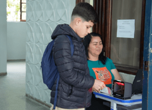 Aluno escaneia carteirinha em leitor de código de barras. Uma pessoa adulta monitora computador próximo ao scanner