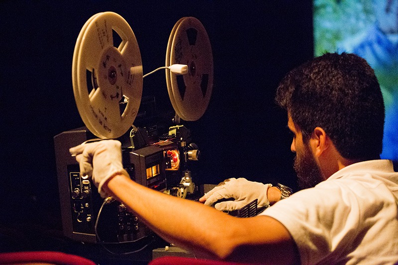 Uma pessoa de cabelo curto manuseia um projetor de filme. A pessoa veste luvas brancas para manipular o filme