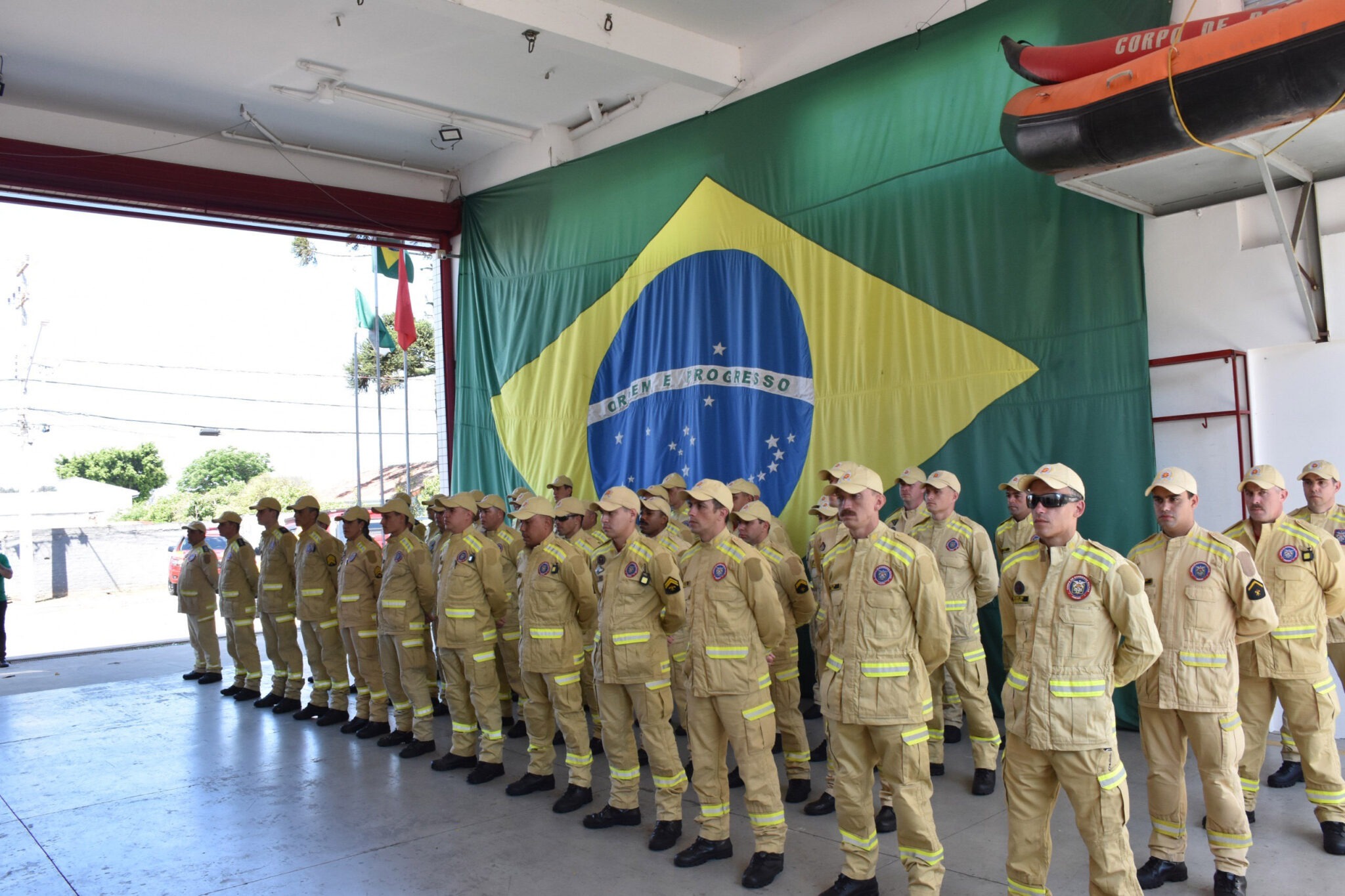 Bombeiros posam em postura de "sentido" em frente à uma bandeira do Brasil estendida no teto.