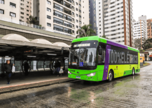 Ônibus de cor verde, estaciona em ponto de ônibus. O clima é chuvoso e na lateral do ônibus é possível ler "100% elétrico"