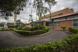 Foto da fachada do hospital Vitória. Uma rua pavimentada faz uma curva vindo de longe da esquerda, curvando próximo à câmera e se distanciando novamente à direita. O hospital é feito de tijolos alaranjados