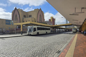 Terminal do Guadalupe. Em primeiro plano, um ônibus da cor branca, estacionado embaixo de um telhado de concreto pintado na cor amarela. Ao fundo, o Santuário de Nossa Senhora de Guadalupe