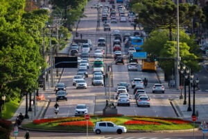 Uma avenida com seis pistas. Vários tipos de veículos circulam em ambas as direções da via