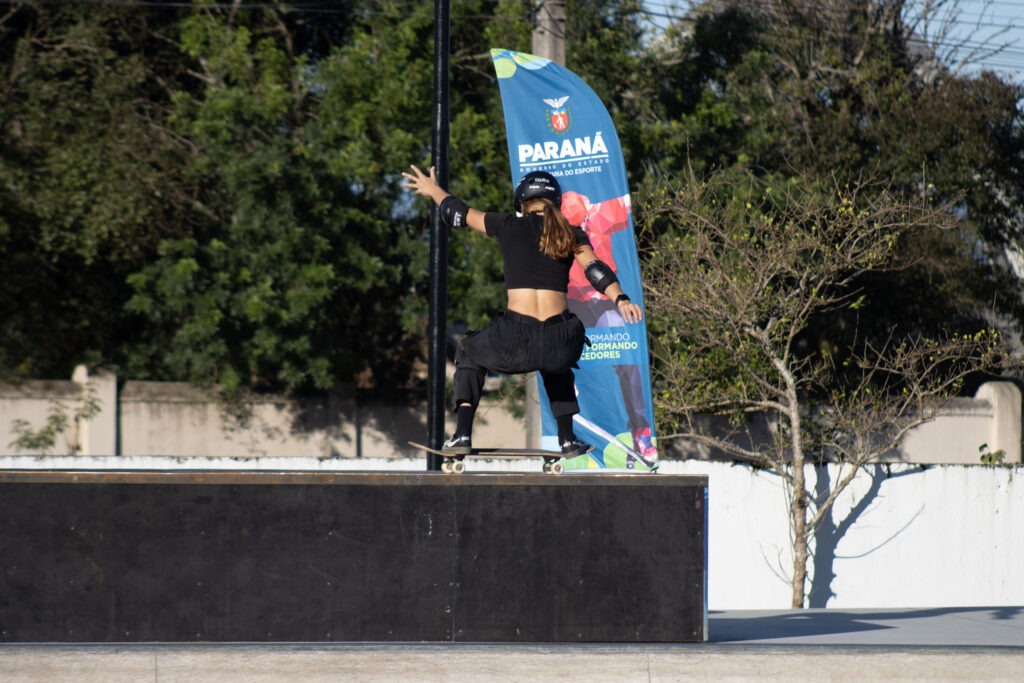 Atleta de cabelo longo faz manobra de skate em estrutura retangular de concreto. A pessoa está em pleno voo, de costas para a câmera.