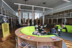 A imagem mostra o interior da Biblioteca Pública do Paraná, um espaço amplo e bem iluminado. O ambiente é moderno, com móveis confortáveis, incluindo um sofá em formato de círculo e cadeiras de design arrojado. As prateleiras de livros se estendem ao longo das paredes, chegando até o nível superior acessível por passarelas. No centro da sala, há um sofá de cor verde-limão e pequenas mesas ao redor, onde algumas pessoas estão lendo. Em primeiro plano, há uma mesa redonda com revistas expostas. Ao lado esquerdo, um grande banner mostra o rosto de um homem sorridente com óculos de armação preta e uma citação: "Não é apenas uma biblioteca. É uma nave especial que te levará aos confins do universo, uma máquina do tempo que te leva do mais recôndito passado ao futuro distante". O local possui colunas brancas, algumas decoradas com desenhos coloridos. Várias pessoas estão sentadas, lendo e aproveitando o espaço em uma atmosfera de tranquilidade e concentração.