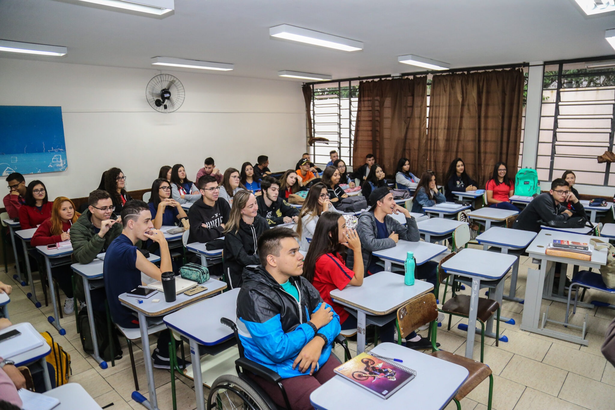 Interior de sala de aula, todas as carteiras estão ocupadas por alunos