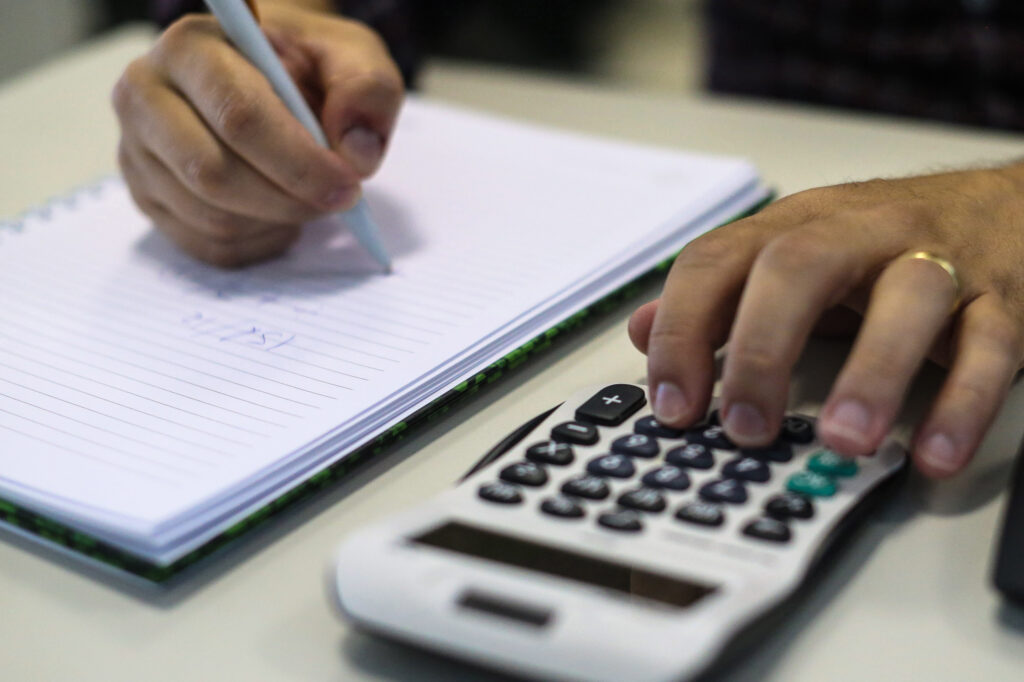 Um par de mãos escreve em caderno e usa calculadora ao lado