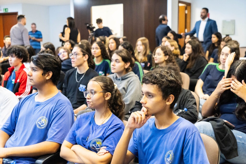 Jovens sentados, vestindo uniforme azul, em cadeiras enfileiradas observam algo para o lado esquerdo da foto