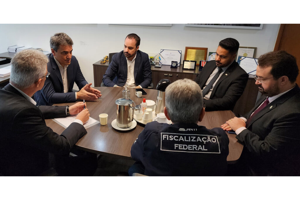 Seis conversam sentados à mesa de madeira. No centro da mesa, uma garrafa térmica e açúcar. Uma das pessoas, virada de costas para a câmera, veste uma camisa com "FISCALIZAÇÃO FEDERAL" escrita nas costas