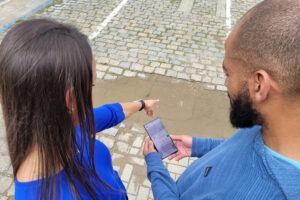 Duas pessoas vestindo azul observam poça de água em calçada. A pessoa à esquerda da imagem aponta para a poça no chão. A pessoa à direita, manipula o celular.