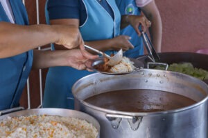 Pessoa vestindo guarda-pó azul serve feijão em prato com arroz