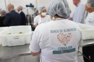 Várias pessoas em cozinha manipulam comida. Todas estão vestindo tocas higiênicas. Nas costas da camiseta de uma das pessoas está escrito "Banco de alimentos, Comida boa"