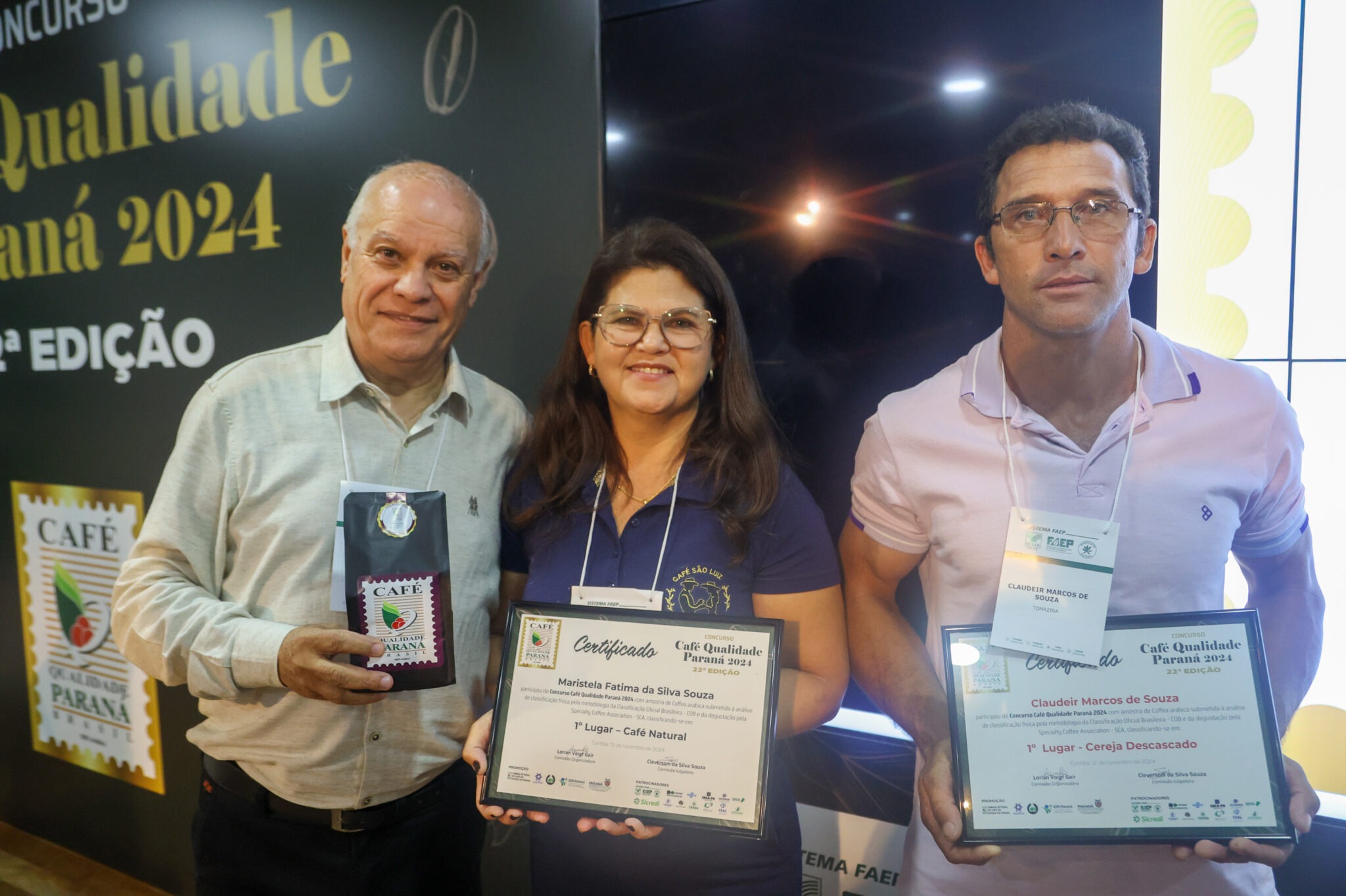 Três pessoam posam para foto. Na esquerda, um homem com camisa de botão segura um pacote de café. No meio, ma mulher de cabelos escuros e óculos, segura um certificado escrito "Primeiro lugar - café Natural" e o nome Maristela Fátima da Silva Souza. Na direita, um homem de cabelo curto, vestindo óculos e camisa polo rosa segura um certificado escrito "Primeiro lugar - Cereja descascado" e o nome Claudeir Marcos de Souza.