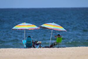 Em primeiro plano, duas pessoas sentadas em cadeiras de praia embaixo de um par de guarda sol. Ao fundo, o mar.