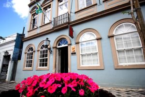 A imagem mostra a fachada do Museu Casa Alfredo Andersen, um edifício histórico de dois andares pintado em azul claro, com detalhes em bege ao redor das janelas e portas. O prédio tem janelas arqueadas no térreo e janelas retangulares no andar superior. Há uma placa vermelha pendurada, indicando o nome do museu, e uma bandeira do Brasil hasteada na parte superior esquerda. Em frente ao edifício, há uma jardineira cheia de flores cor-de-rosa vibrantes, adicionando um toque de cor ao cenário. A fachada do museu está bem conservada e em estilo clássico, com uma aparência acolhedora e charmosa.