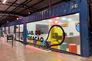 Fachada de ambiente de inovação. O ambiente foi construído dentro de um contêiner azul. Uma das paredes é feita de vidro e foi adesivado com um ícone de lâmpada e o nome "LAB DE IDEAÇÃO"