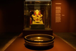 A imagem mostra uma exposição de arte asiática no Museu Oscar Niemeyer (MON). Em destaque, há uma estátua dourada de Ganesha, uma divindade hindu com cabeça de elefante, exposta dentro de uma vitrine de vidro iluminada, posicionada no centro da sala. A estátua repousa sobre uma base elevada e está cercada por luzes que realçam seus detalhes e cores vibrantes. À frente da vitrine, sobre uma mesa, há um grande recipiente redondo de metal contendo pequenas esferas brancas, sugerindo algum tipo de objeto ritual ou decorativo. A parede ao fundo tem um texto explicativo com o título "Os Mauryas", descrevendo a temática da exposição em letras brancas sobre um fundo marrom. O ambiente possui iluminação baixa, criando um clima de contemplação.