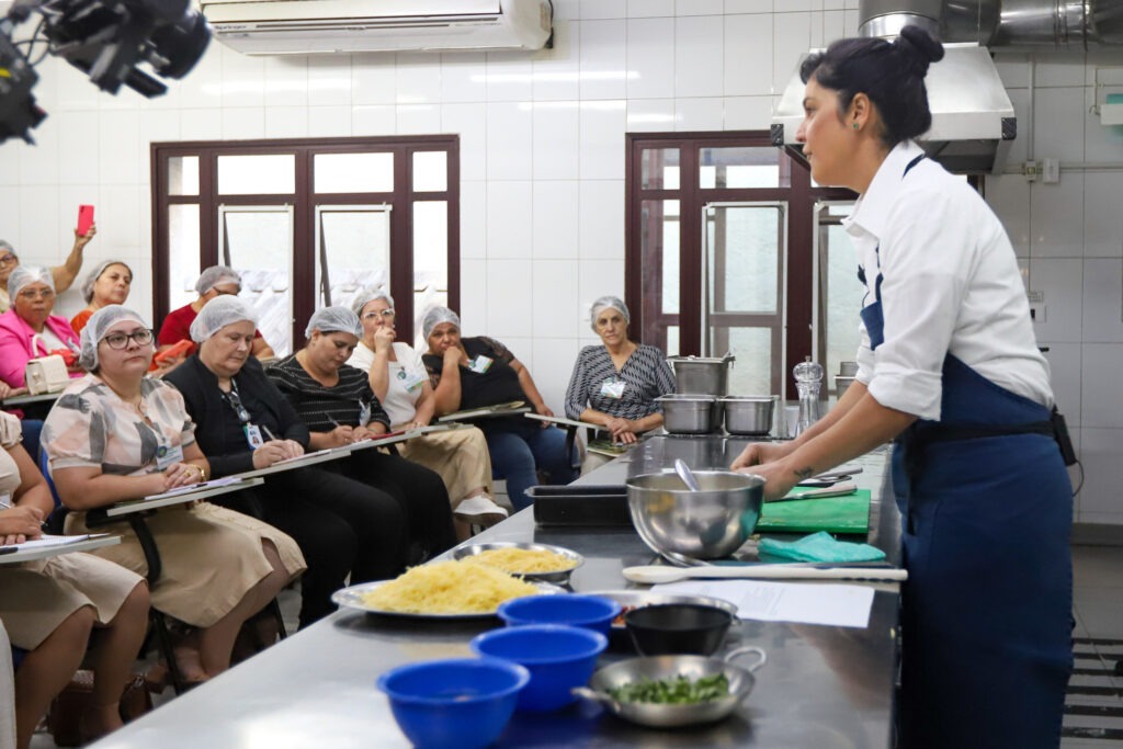 A imagem mostra um ambiente de cozinha industrial onde a chef de cozinha Manu Buffara está em pé, de perfil, usando um avental azul sobre uma camisa branca. Ela está em frente a uma bancada de metal com ingredientes e utensílios de cozinha, como tigelas, pratos com alimentos e uma tábua de corte. À sua frente, várias mulheres estão sentadas em cadeiras, vestindo uniformes bege e toucas descartáveis de proteção no cabelo. Elas parecem atentas, com algumas fazendo anotações em pranchetas. Ao fundo, há uma parede de azulejos brancos e janelas de madeira. O clima é de aprendizado e interação, sugerindo uma aula ou oficina prática.