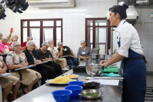 A imagem mostra um ambiente de cozinha industrial onde a chef de cozinha Manu Buffara está em pé, de perfil, usando um avental azul sobre uma camisa branca. Ela está em frente a uma bancada de metal com ingredientes e utensílios de cozinha, como tigelas, pratos com alimentos e uma tábua de corte. À sua frente, várias mulheres estão sentadas em cadeiras, vestindo uniformes bege e toucas descartáveis de proteção no cabelo. Elas parecem atentas, com algumas fazendo anotações em pranchetas. Ao fundo, há uma parede de azulejos brancos e janelas de madeira. O clima é de aprendizado e interação, sugerindo uma aula ou oficina prática.