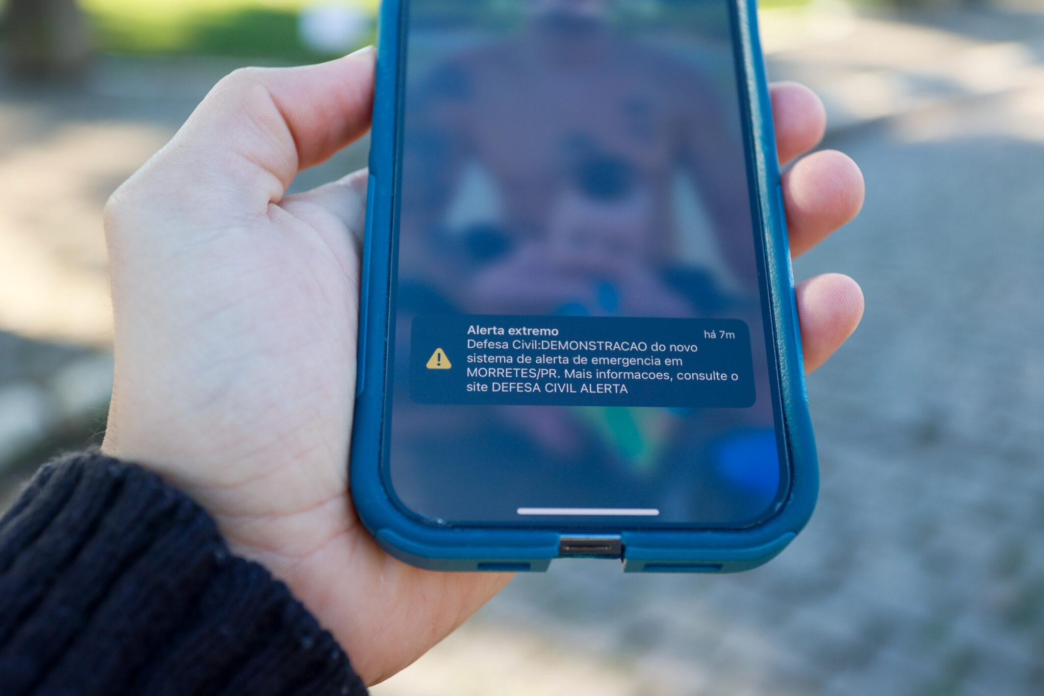 Mão esquerda segura celular. Na tela, um aviso: "Alerta extremo"
