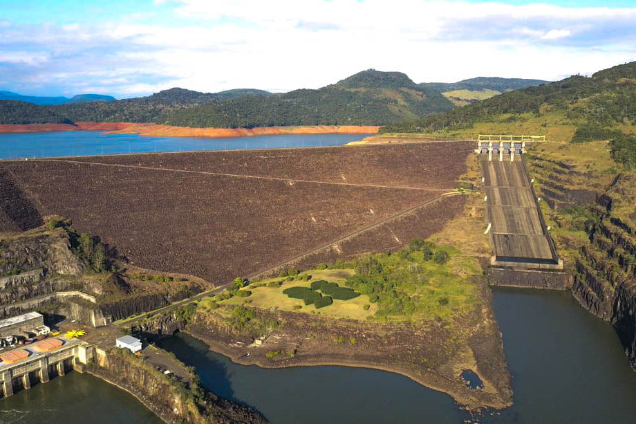 A imagem mostra uma vista aérea da Usina Hidrelétrica Foz da Areia, localizada em um cenário montanhoso e verdejante. No centro, destaca-se o enorme paredão da barragem revestido com pedras escuras, que retém as águas de um grande reservatório azul à esquerda. À direita, estão as estruturas da vertedoura, com uma longa rampa de concreto inclinada, usada para liberar o excesso de água do reservatório. Abaixo da barragem, vê-se o rio e parte da infraestrutura da usina, incluindo edifícios e turbinas. O ambiente ao redor é cercado por montanhas cobertas de vegetação e céu azul com algumas nuvens.