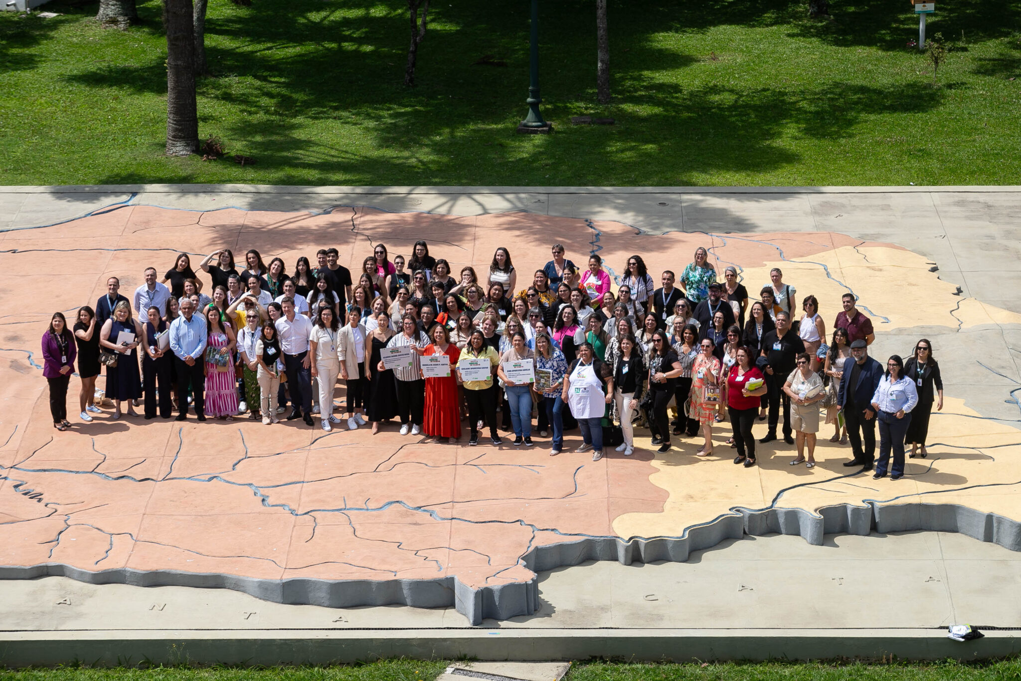 A imagem apresenta um grande grupo de pessoas, destacando as merendeiras finalistas do concurso "Melhor Merenda do Paraná". Elas estão reunidas sobre um enorme mapa em relevo do estado do Paraná, localizado em uma praça ao ar livre. O grupo inclui mulheres e homens. Todos posam sorrindo para a foto. Ao fundo, o gramado e as árvores reforçam o ambiente externo iluminado por luz natural.
