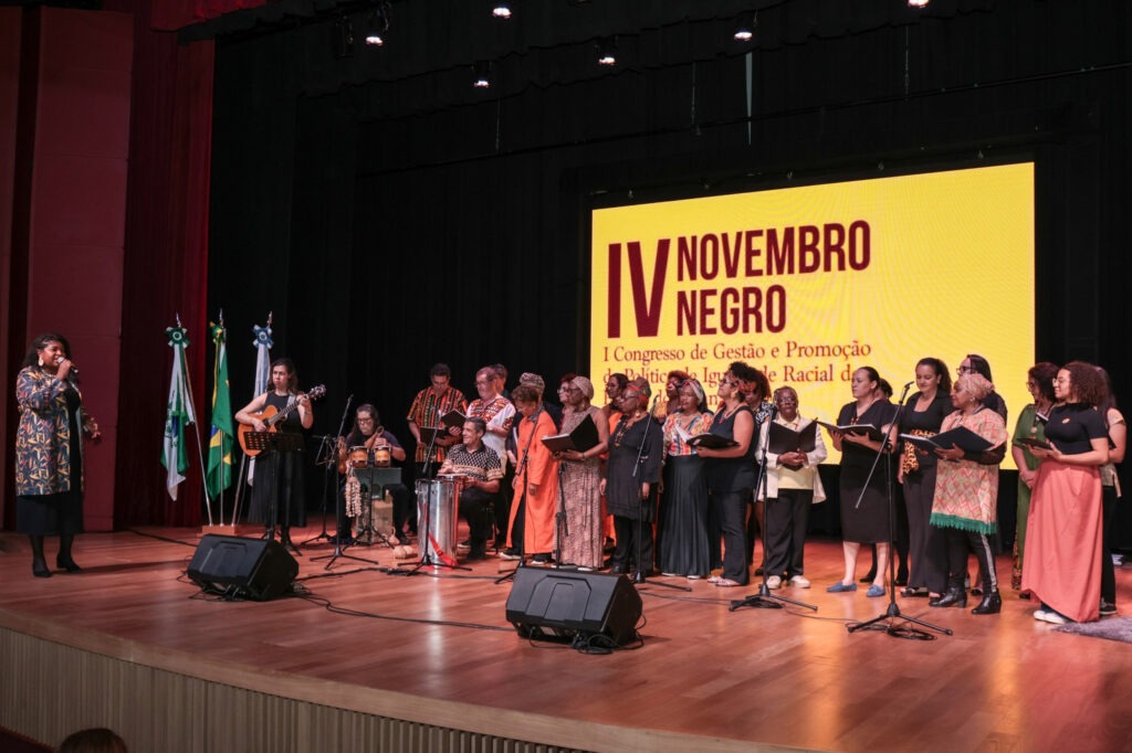 Várias pessoas de pele escura em palco de teatro. Ao fundo, um telão de fundo amarelo com escrita em vermelho escuro: IV Vivendo Negro