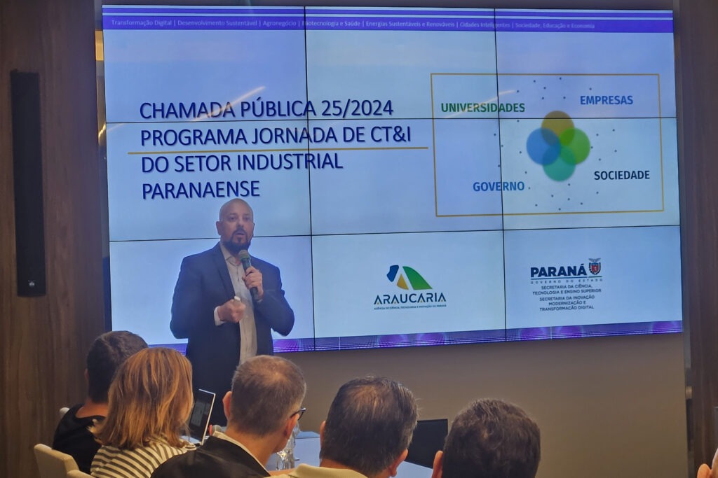 Uma pessoa vestindo terno azul e segurando um microfone está em frente de um telão feito de nove telas juntas. Na tela: "Chamada pública programa jornada de CT&I do setor industrial paranaense"