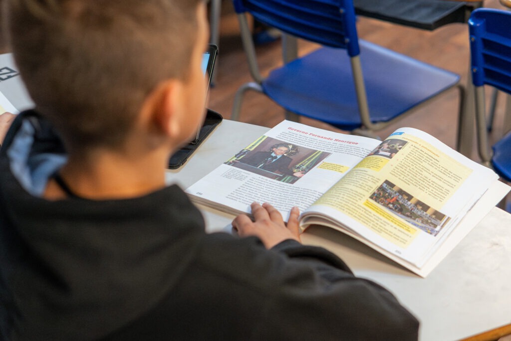 A imagem mostra um estudante em sala de aula, concentrado em um livro didático aberto sobre a mesa. A página exibe textos e imagens relacionados a conteúdo escolar, sugerindo um momento de leitura ou estudo. No plano de fundo, é possível notar cadeiras azuis, características do ambiente educacional. A cena enfatiza a rotina de aprendizado dos alunos e o ambiente organizado da escola.