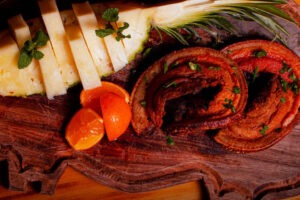 Foto de tábua de madeira com tiras de queijo, pedaços de laranja fatiada e rodelas de carne de porco com temperos