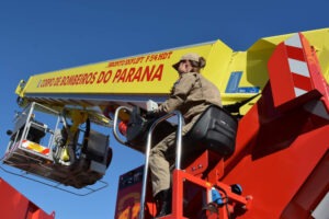 A imagem mostra uma bombeira do Corpo de Bombeiros do Paraná operando um veículo especial equipado com uma plataforma elevatória articulada, identificada como Bronto Skylift F54 HDT. O equipamento é pintado nas cores vermelho e amarelo, com o nome "Corpo de Bombeiros do Paraná" destacado na estrutura. A bombeira veste o uniforme oficial bege e está sentada na cabine de comando do equipamento, que está elevado. O céu ao fundo está limpo, com um tom azul vibrante, sem nuvens.