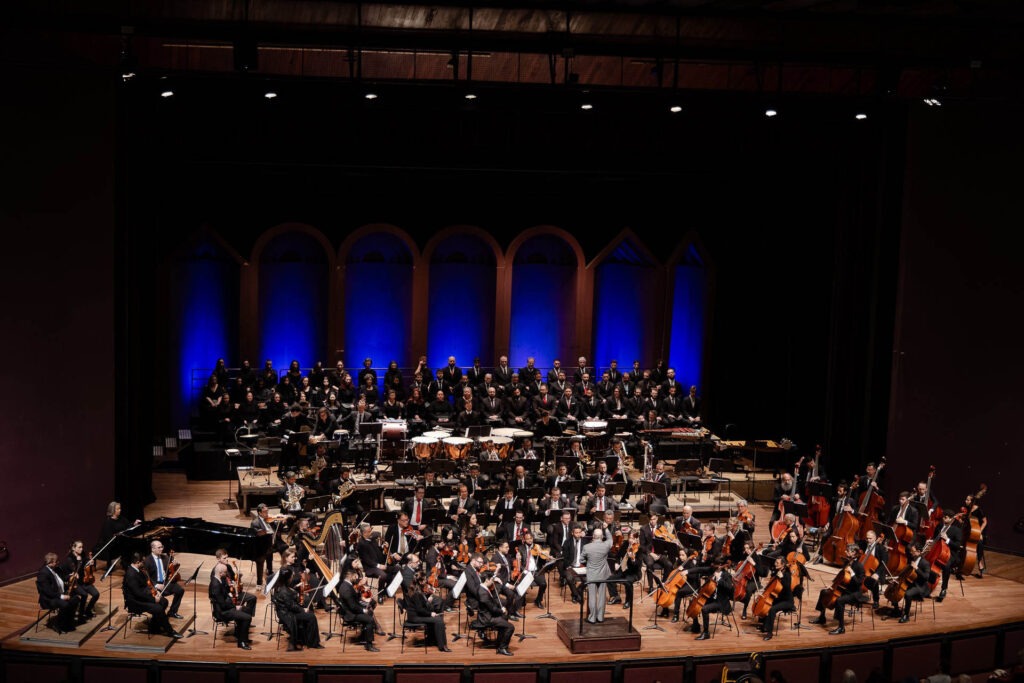 A imagem mostra a Orquestra Sinfônica do Paraná durante uma apresentação em um palco de teatro. O grupo ocupa toda a área central do palco, com músicos posicionados em semicírculo na parte frontal e o coro ao fundo, em uma formação elevada. O maestro, em traje formal cinza, está de pé em um pequeno pódio central, conduzindo a performance. Os músicos tocam uma variedade de instrumentos, incluindo violinos, violoncelos, contrabaixos, instrumentos de sopro e percussão, além de uma harpa. O coro, composto por várias pessoas vestidas em trajes pretos, está disposto em três níveis de arquibancadas no fundo do palco. A iluminação destaca o fundo azul e arcos decorativos em tons dourados, criando uma atmosfera elegante. A plateia não é visível, mas os assentos do auditório ocupam o primeiro plano da imagem.