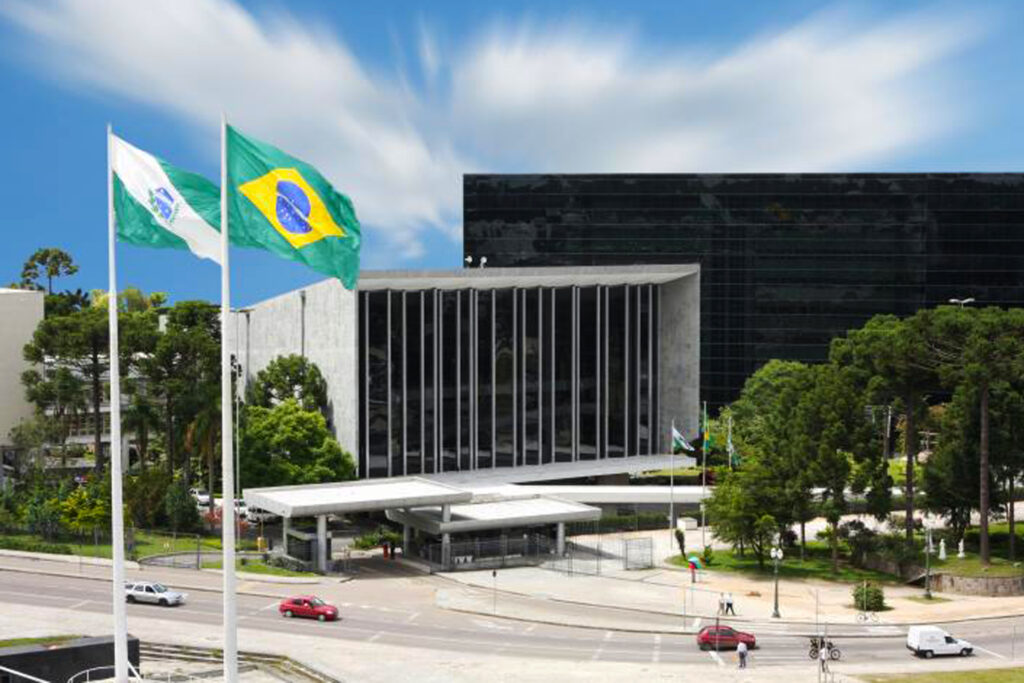 A imagem mostra a fachada da Assembleia Legislativa do Paraná (ALEP), localizada em Curitiba. No primeiro plano, estão hasteadas duas bandeiras em mastros altos: a bandeira do Brasil à direita e a bandeira do estado do Paraná à esquerda, ambas balançando ao vento. O edifício da ALEP aparece ao fundo, com uma arquitetura moderna que combina concreto e vidro, destacando uma grande área envidraçada com pilares verticais. Em frente ao prédio, há uma via asfaltada por onde passam alguns carros, bicicletas e pedestres. Árvores e áreas verdes cercam o local, complementando a paisagem urbana. O céu está claro com algumas nuvens brancas.