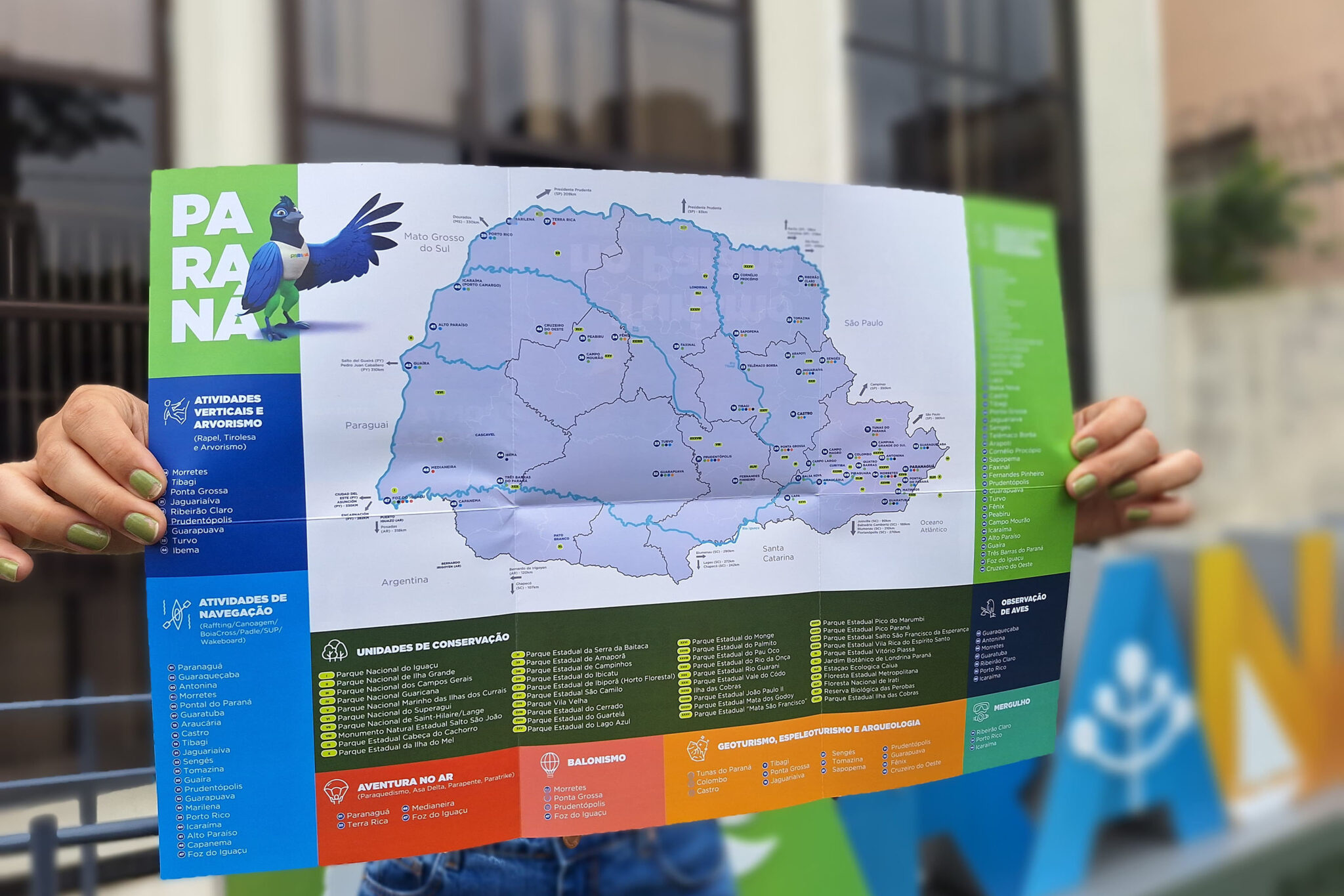 A imagem apresenta um mapa turístico do Paraná, produzido pela Secretaria de Turismo do estado, sendo segurado por uma pessoa com esmalte verde nas unhas. O material destaca atrações de ecoturismo e aventura, incluindo atividades verticais, arvorismo, balonismo, mergulho e observação de aves. As áreas protegidas, como parques nacionais e estaduais, estão marcadas no mapa, que usa cores vibrantes para diferenciar categorias de atividades. À esquerda, um personagem ilustrado de ave simboliza a biodiversidade do estado.