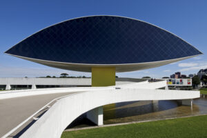 A imagem retrata o Museu Oscar Niemeyer (MON), em Curitiba, com destaque para a estrutura icônica em formato de "olho". O edifício principal é sustentado por uma base amarela com um desenho preto que lembra traços de um olho, refletido em um espelho d'água ao redor. A parte superior possui uma superfície envidraçada preta com padrão de losangos, que reflete o céu e edifícios ao fundo. À direita, uma rampa curva branca conecta o prédio ao solo, enquanto o gramado ao redor e algumas árvores ao fundo compõem a paisagem. O céu está claro, com tonalidades de azul, indicando um dia ensolarado.