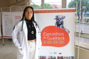 A imagem mostra Ádana Garigsãnh Bernardo, estudante indígena Kaingang, que está no segundo ano de Medicina na Unioeste. Ela veste um jaleco branco e um estetoscópio em volta do pescoço, com um colar e brincos que remetem à cultura indígena. Ao fundo, um banner com a inscrição "Caminhos de Guairacá" exibe a imagem de um busto de uma figura histórica indígena e detalhes gráficos com padrões tribais. A cena ocorre em um ambiente interno iluminado por luz natural, com mapas e informações culturais expostas em painéis.