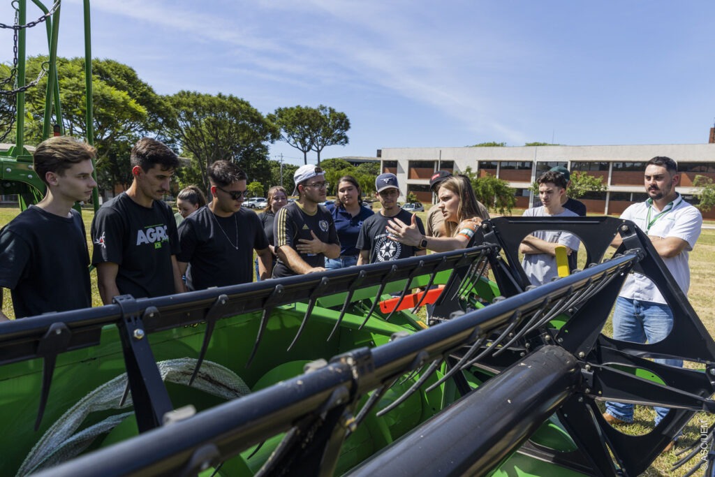 A imagem retrata um grupo de estudantes observando e recebendo explicações sobre uma máquina agrícola de grande porte, destacada em primeiro plano. Uma mulher, aparentemente instrutora, gesticula enquanto apresenta detalhes técnicos do equipamento.