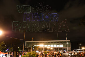 Drones luminosos formam as palavras "Verão Maior Paraná". Os drones estão sobrevoando o Palácio Iguaçu a noite.