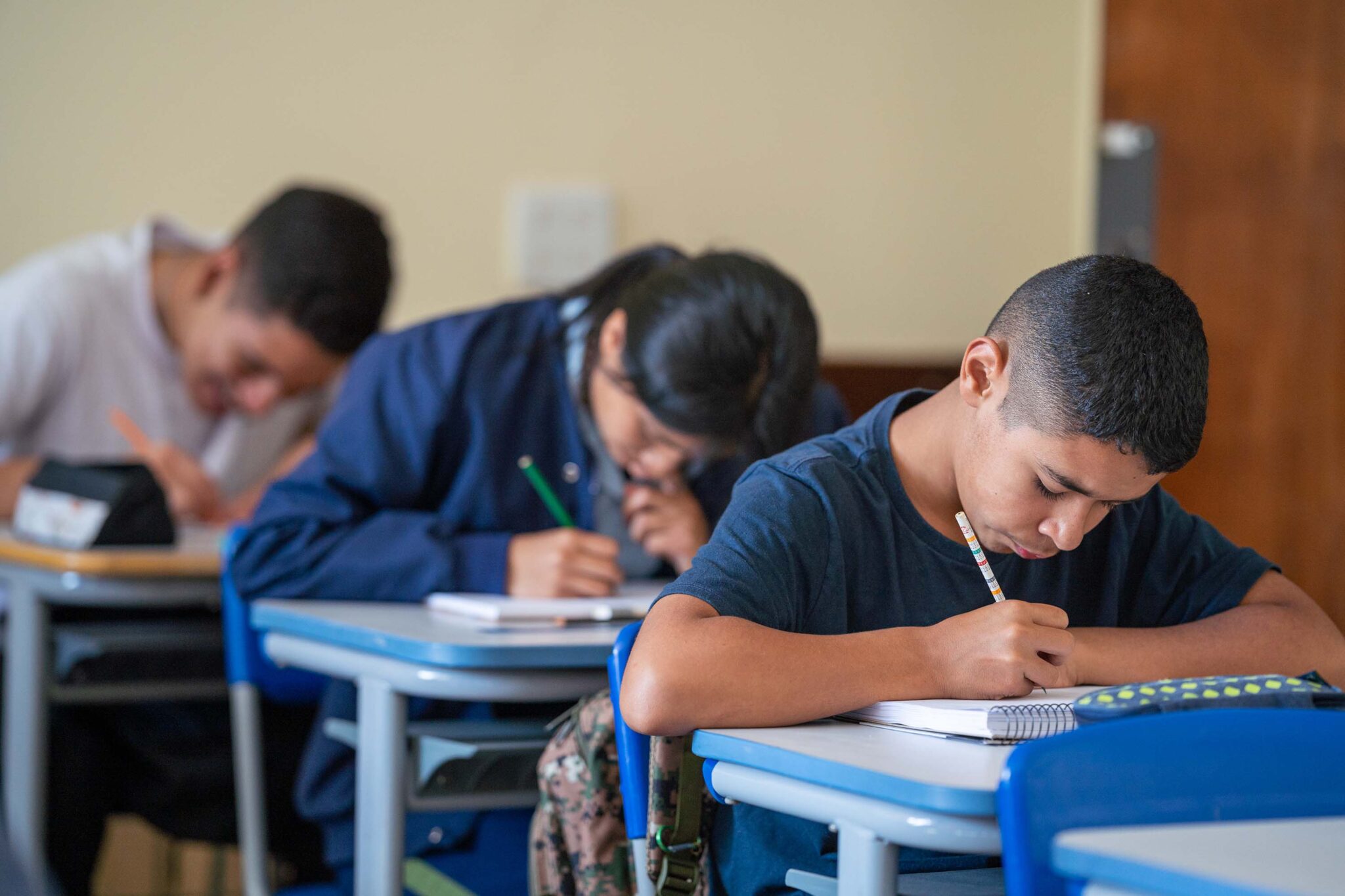 Alunos realizando atividade. As mesas e cadeiras onde os alunos estão sentados estão alinhadas.