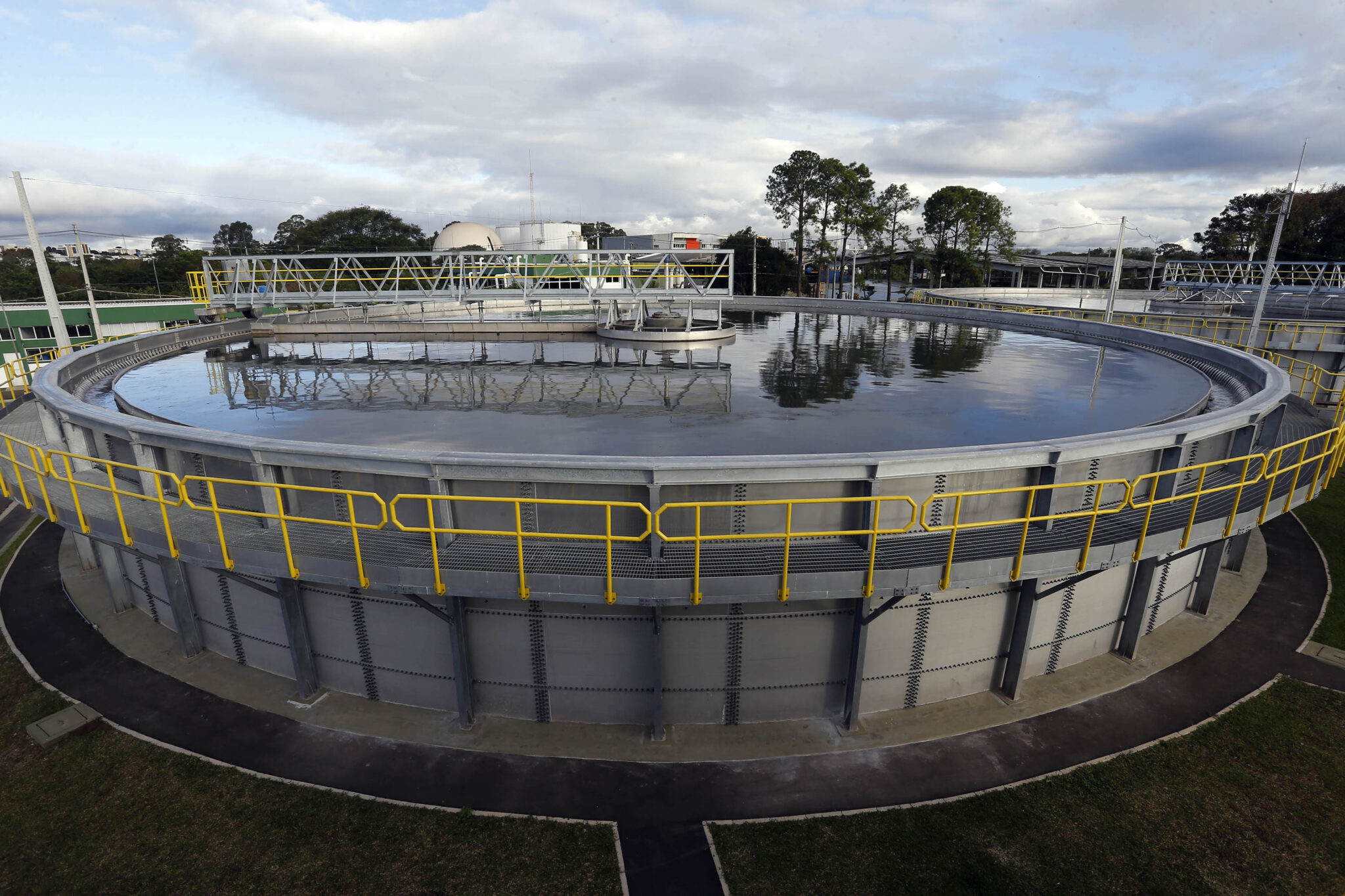 A imagem mostra uma estação de tratamento de água ou esgoto com um grande tanque circular de sedimentação, feito de concreto e cercado por grades de segurança pintadas de amarelo. O tanque está cheio de água refletindo parte da estrutura ao redor. Ao fundo, é possível observar outras instalações da estação, árvores e uma área com céu parcialmente nublado