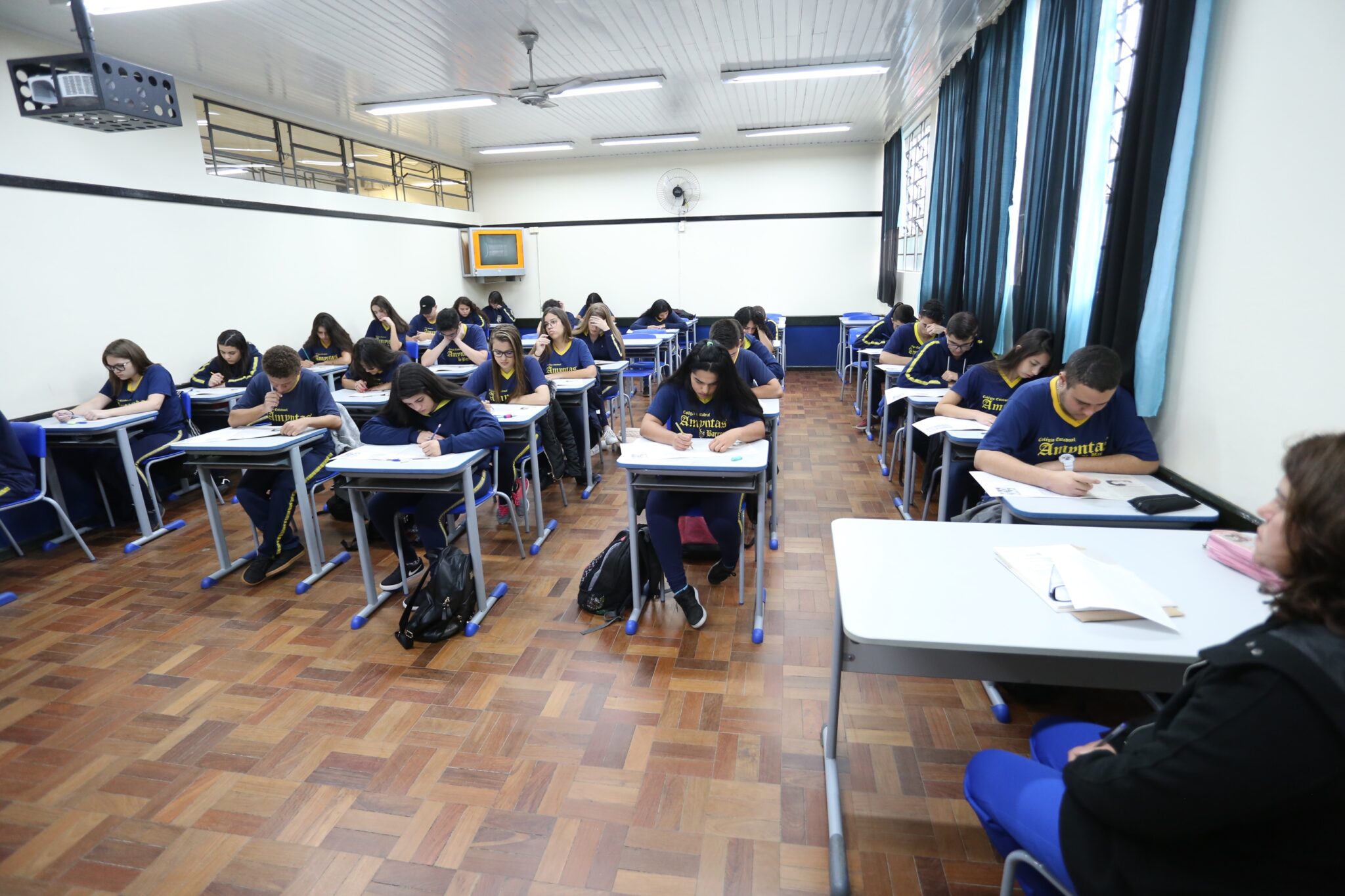 A imagem mostra uma sala de aula com diversos estudantes uniformizados, concentrados em suas mesas enquanto realizam alguma atividade escrita. As carteiras estão organizadas em fileiras, e os alunos parecem estar em um ambiente de prova ou estudo supervisionado. Ao fundo, há um ventilador de parede e uma televisão desativada presa ao suporte. A sala é iluminada por luzes artificiais no teto e pela claridade vinda das janelas com cortinas azuis. O piso é de madeira e uma pessoa, possivelmente a professora, está sentada no canto direito, supervisionando os estudantes.