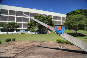 Em primeiro plano, uma escultura de concreto imitando um ponteiro com o símbolo da Unioeste em sua metade. Ao fundo, um prédio de três andares atravessa a imagem de ponta a ponta.